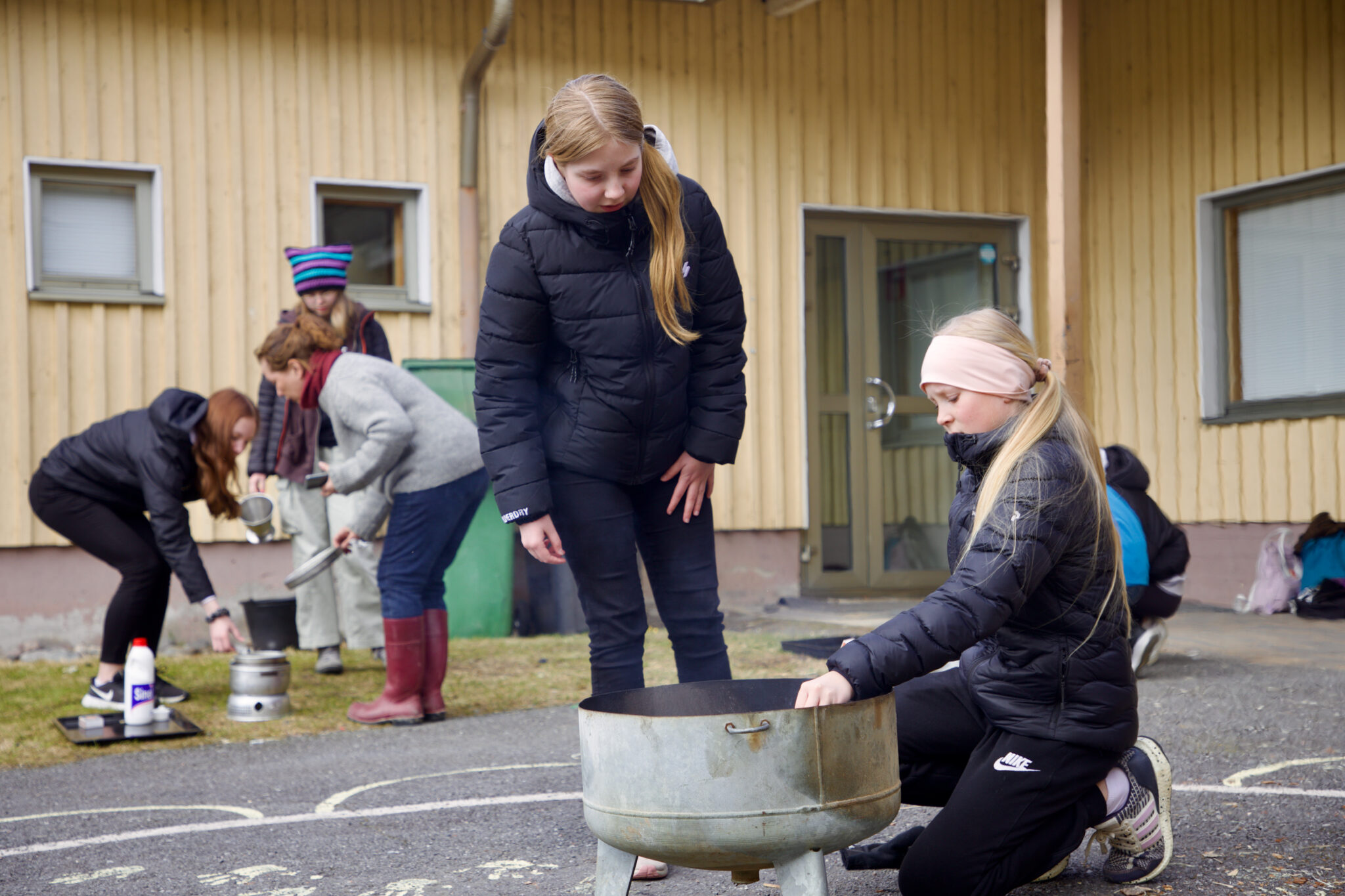 Erätaitopisteellä Aija Koivisto (oik.) ja Helmiina Kytölä sytyttivät nuotiotulta koulun sisäpihalla. Taustalla 4H:n Hanna Honkaniemi ohjaa Aada Kuoppa-ahoa ja Isabella Lassilaa trangian käytössä.