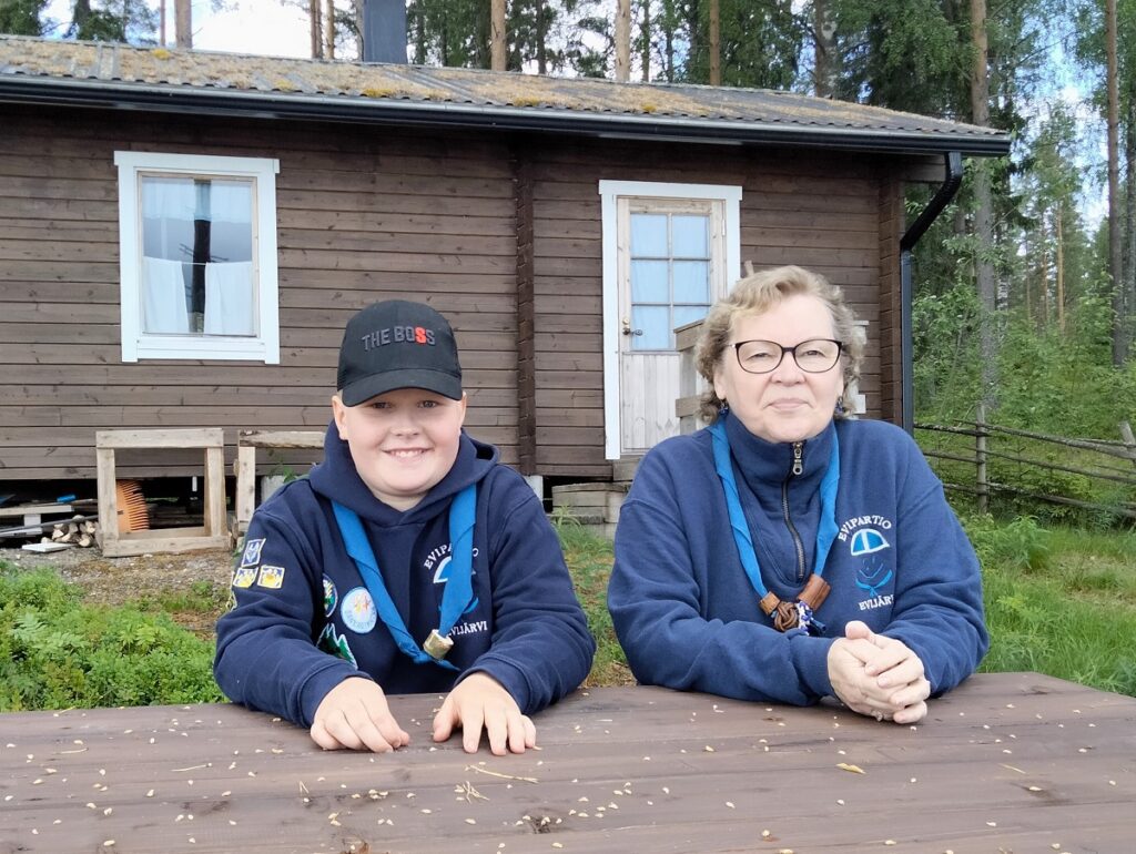 Eetu Koskipuisto ja Maritta Hanhikoski ovat molemmat innokkaita partiolaisia. Eetu kuuluu Evipartion Seikkailijoihin, Maritta-mummo liittyi partioon jo vuonna 1966. Taustalla Korkeamaan partiomaja.